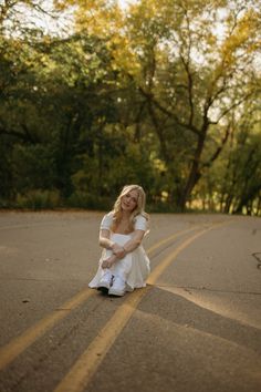 a woman sitting on the side of a road wearing a white dress and holding a purse