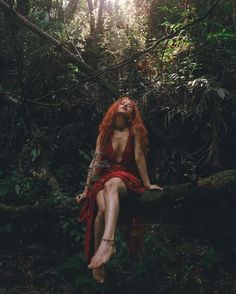 a woman with red hair is sitting on a log in the woods and looking up