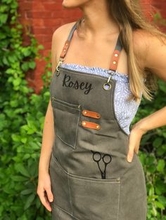 a woman wearing an apron with scissors drawn on the front and side pockets, standing in front of a brick wall