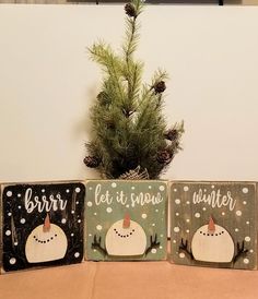 three wooden signs with snowmen and pine cones on them sitting next to a christmas tree