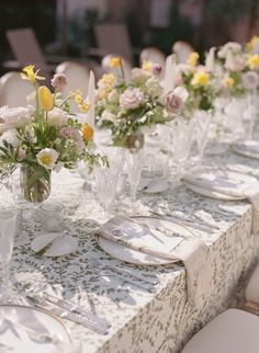 a long table is set with white and yellow flowers in vases, plates and silverware