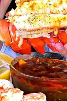 a person holding up a large lobster sandwich over a bowl of dipping sauce and lemon wedges