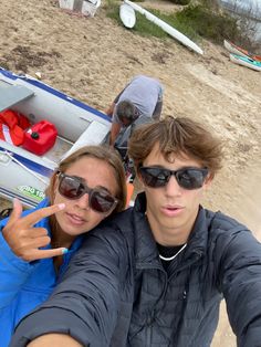 two people taking a selfie in front of a small boat on the beach with one pointing at the camera