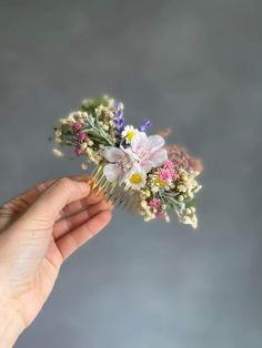 a hand holding a comb with flowers and leaves on it in front of a gray background