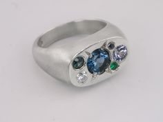 Journey back to an era of opulence with this candy studded cushion signet ring. At its centre, a mesmerising 6mm London blue topaz captures the heart of celestial beauty, surrounded by a constellation of gems: 4mm lilac grey spinel, 3mm Australian teal sapphire, 3mm moissanite, 2mm lab emerald, and a 1.9mm salt and pepper diamond. Each gem is thoughtfully chosen to create a harmonious blend of colours and light, echoing the ancient world's fascination with the cosmos. This ring not only celebrat Celestial Oval Signet Ring For Anniversary, Celestial Oval Multi-stone Ring, Teal Sapphire, Lilac Grey, Pepper Diamond, Pearl Gemstone, Black Rhodium, Salt And Pepper Diamond, Raw Gemstones