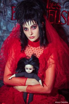 a woman in red dress holding a doll with black hair and makeup on her face