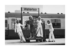 four women standing in front of a train at a station with their arms around each other