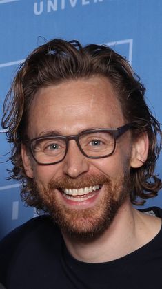 a man with long hair and glasses smiling in front of a blue backdrop at an event