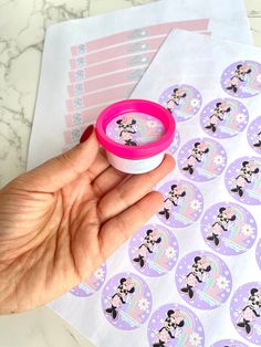 a hand holding a pink cup with minnie mouse stickers on it next to some envelopes