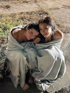 two women wrapped in blankets sitting on the ground next to each other and looking at the camera