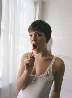 a woman brushing her teeth in front of a window with white curtains and brick walls