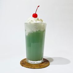 a green drink with whipped cream and a cherry on the top sits on a woven coaster