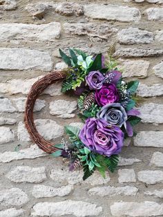 a wreath with purple flowers and greenery hanging on a stone wall