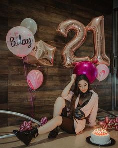 a woman sitting in front of a birthday cake with balloons on the wall behind her