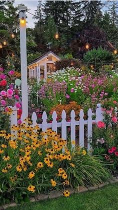 a garden filled with lots of flowers next to a white picket fence covered in lights