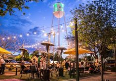 people are sitting at tables under umbrellas in the evening near an outdoor bar and water tower