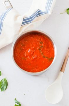 a bowl of tomato soup next to a spoon and napkin on a white surface with basil leaves