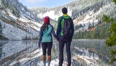 a man and woman holding hands while looking at the mountains reflected in water on a sunny day