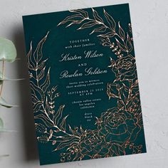 a green wedding card with gold foil flowers and leaves on the front, sitting next to a potted plant