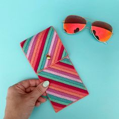 a woman's hand holding a pink and green striped wallet next to sunglasses on a blue background