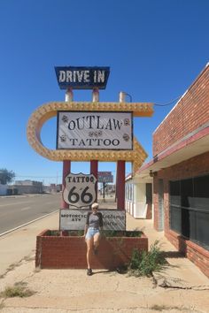 a woman is standing in front of a sign for a tattoo parlor that says drive in