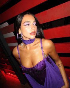 a woman with purple makeup is posing in front of a red wall and wearing a purple dress