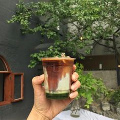 a person holding up a cup in front of a building with trees on the other side