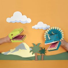 two hands are holding paper dinosaurs in front of a mountain and palm tree on an orange background