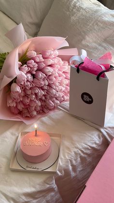 a pink birthday cake sitting on top of a bed next to a bag and flowers