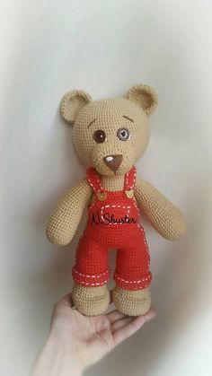 a hand holding a small teddy bear in red overalls