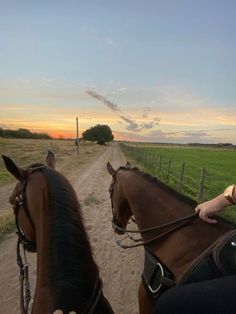 two people riding horses on a dirt road at sunset or dawn with grass and trees in the background