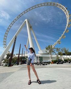 a woman standing in front of a large ferris wheel on a sunny day with her hand up