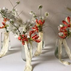 three glass vases with flowers in them on a white tableclothed surface, one is empty and the other has ribbons tied around it