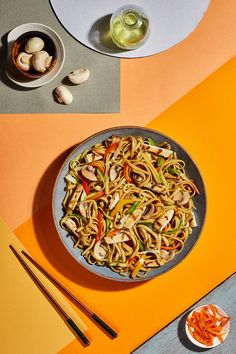 a plate of noodles with mushrooms and chopsticks on an orange tablecloth next to a bowl of vegetables