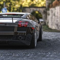 the rear end of a black sports car on a cobblestone road with trees in the background