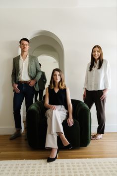 three people are standing and sitting on a green chair in front of a white wall