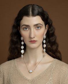 a woman with long dark hair and blue eyes wearing pearls on her necklace, standing in front of a brown background