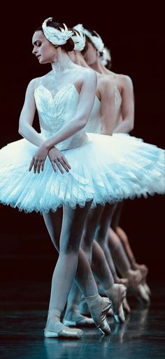 three ballerinas in white tutus and ballet shoes, one is holding the other's leg