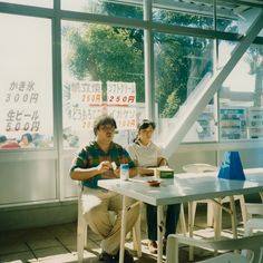 two people sitting at a table in front of a window with signs on it and one person holding a cup