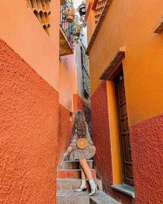 a woman is sitting on the stairs in an alleyway with orange walls and pots hanging above her