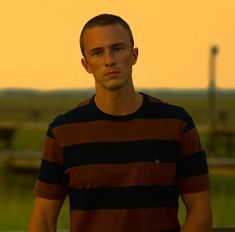 a young man standing in front of a field at sunset with his hands on his hips