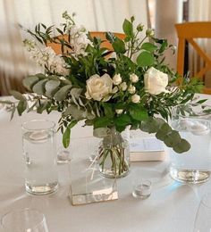 a vase with white flowers and greenery on a table set for an elegant dinner