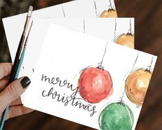 two cards with christmas ornaments on them being held by a woman's hand and holding a pen