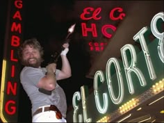 a man with a beard holding a bottle in front of a neon sign that reads gambling