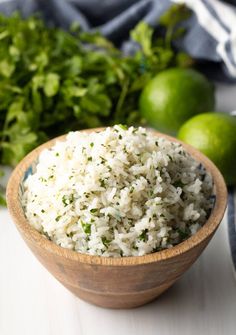 a wooden bowl filled with rice next to limes and cilantro on a white table