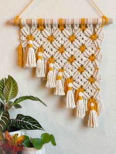 a crocheted wall hanging with tassels and flowers in front of a potted plant