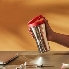 a person holding a cup with the lid open on a table next to a laptop