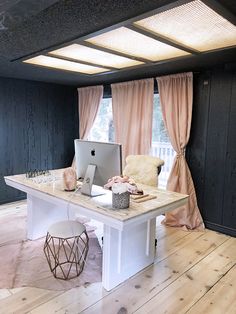 a desk with a computer on top of it in front of a window covered by pink drapes