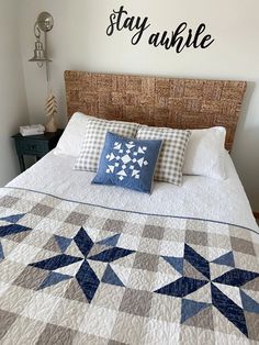 a bed with a blue and white quilt on it's headboard next to a wall that says stay awhile