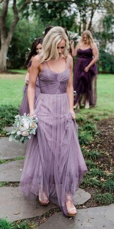 a woman in a long purple dress walking down a path with her bridesmaids